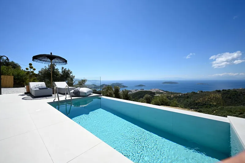 The infinity pool under a black parasol, with the green hillside and the sea and islets beyond